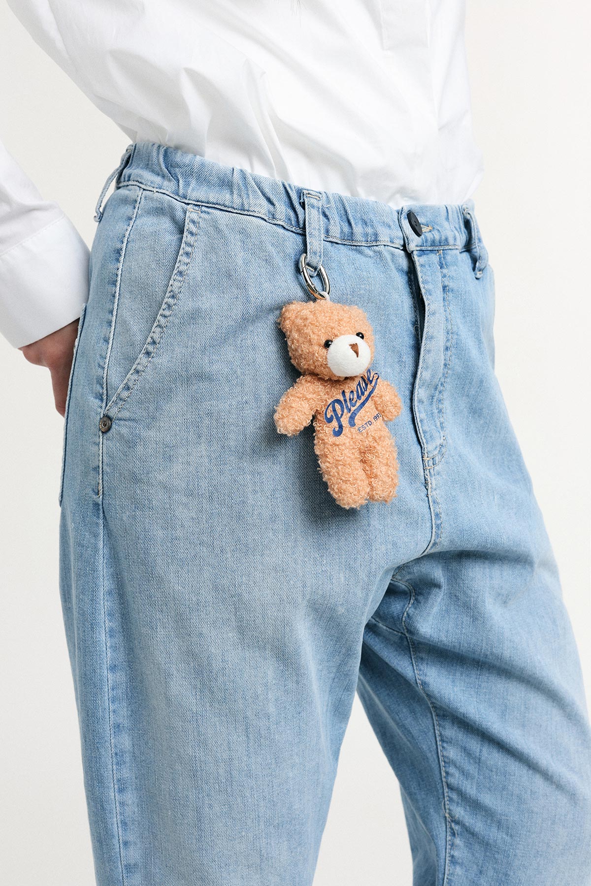 Close-up of small brown teddy bear keychain clipped to light-blue denim jeans pocket on person wearing white shirt