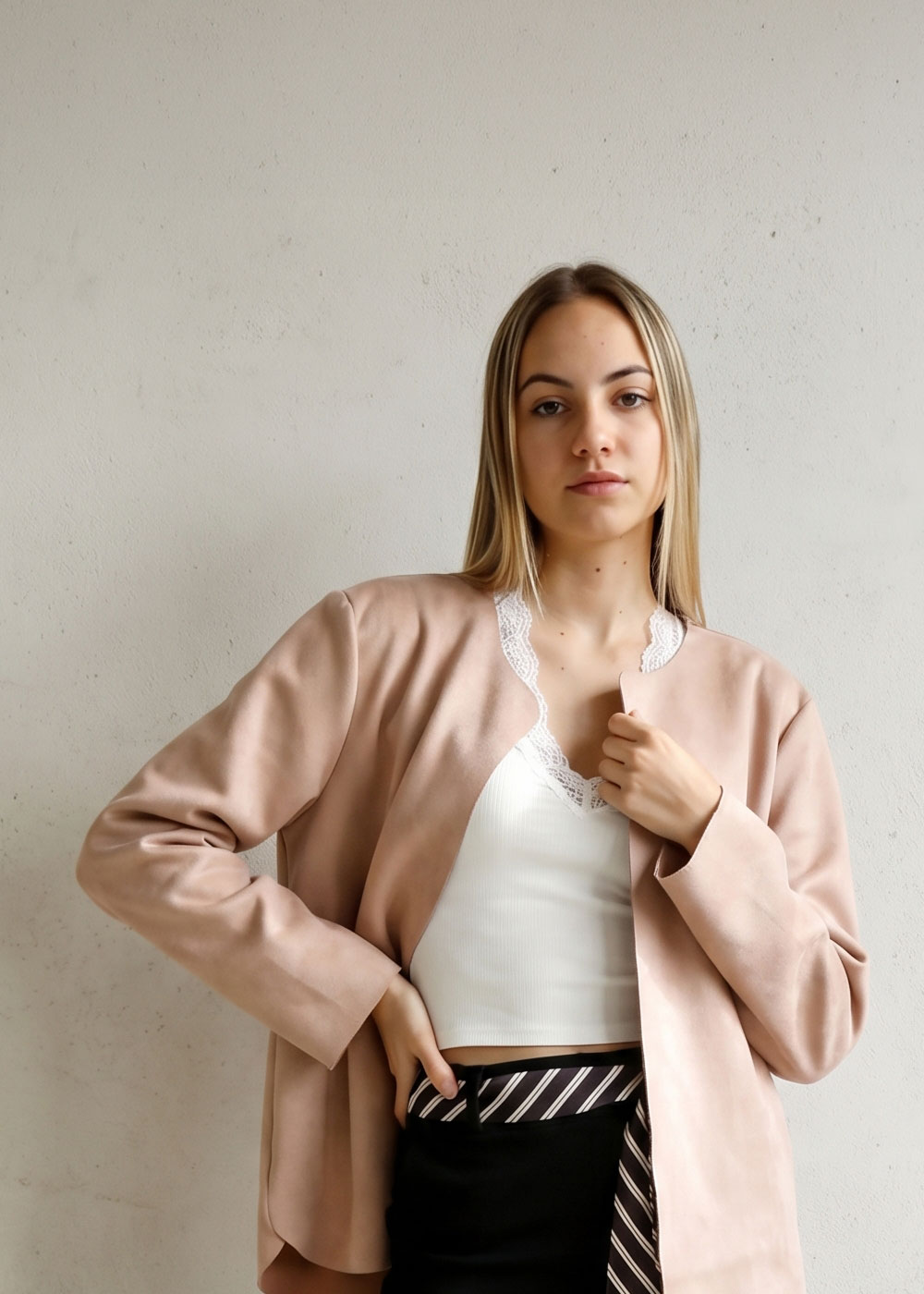 Young woman in pale pink jacket and white lace-trimmed camisole with black skirt and striped belt, neutral pose