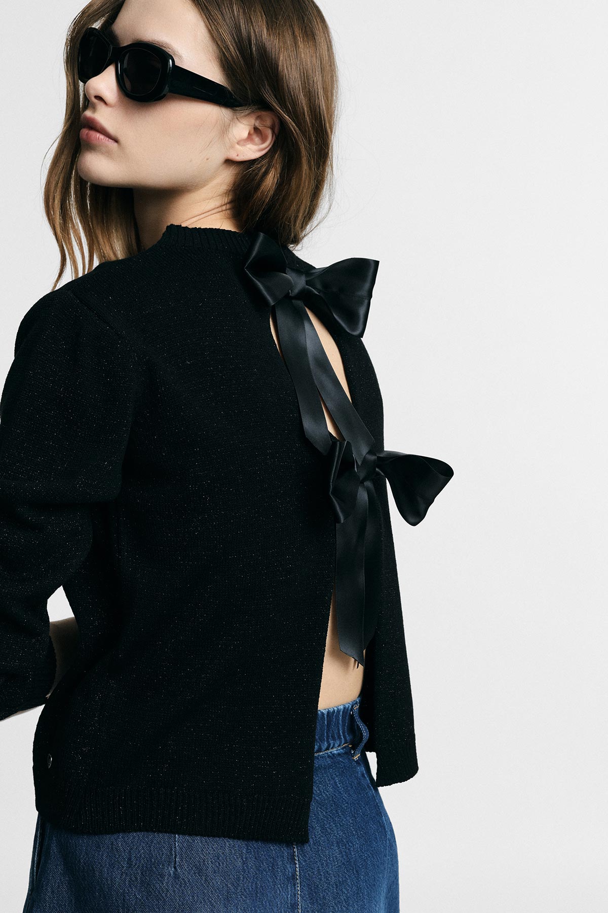 Woman wearing a black open-back sweater tied with two satin bows, dark sunglasses and blue jeans against a white background