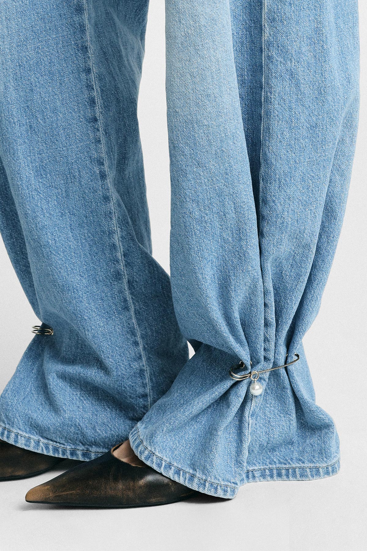 Close-up of light-blue wide-leg jeans cinched at the ankles with decorative safety-pin hoops and a small pearl charm, paired with pointed brown shoes