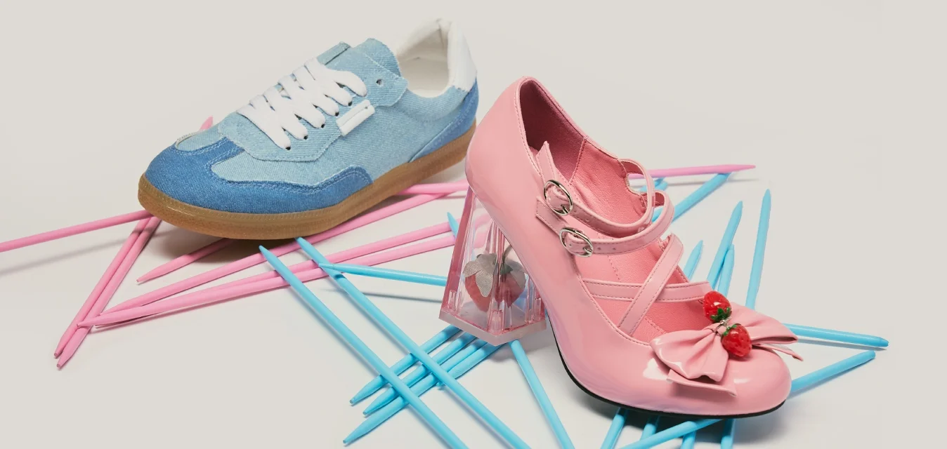 Scarpa con tacco alto rosa con decorazione a ciliegia e sneaker blu posate su bastoncini di plastica rosa e blu sovrapposti su sfondo chiaro.
