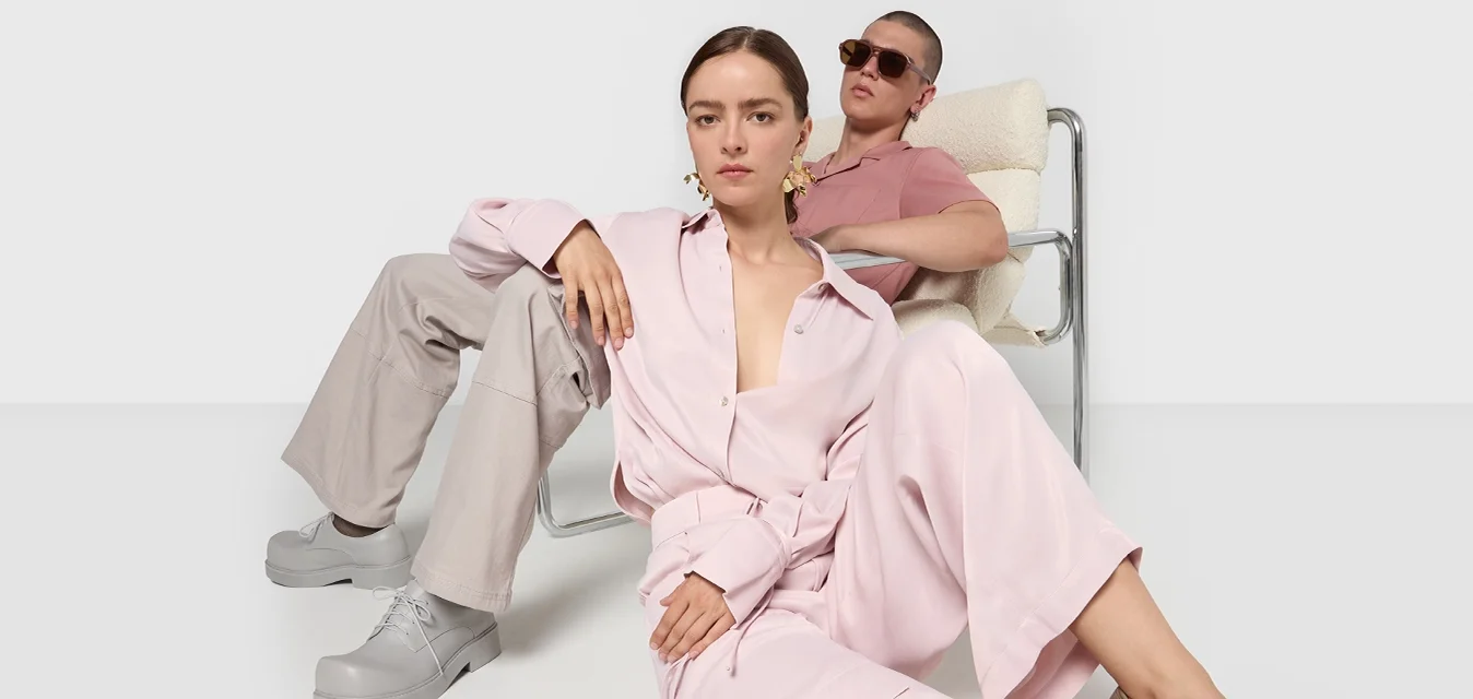 Due persone posano in abbigliamento casual. Uno indossa una camicia button-up rosa chiaro e pantaloni larghi abbinati; l'altro ha una camicia rosa e pantaloni beige.