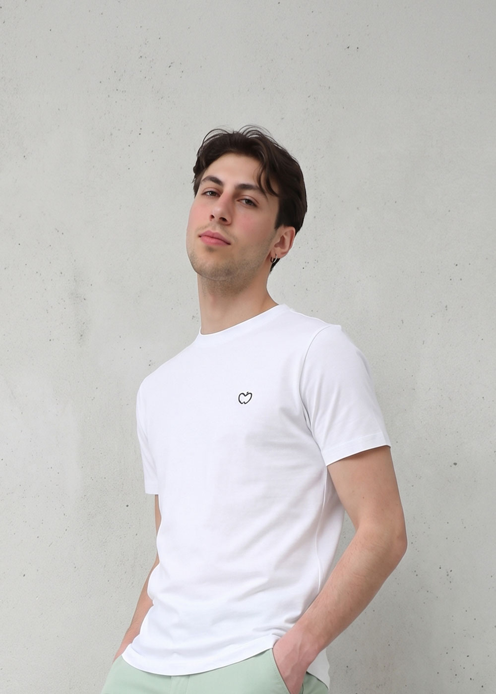 Young man wearing a white T-shirt with a small heart logo, standing against a pale concrete wall with hands in pockets