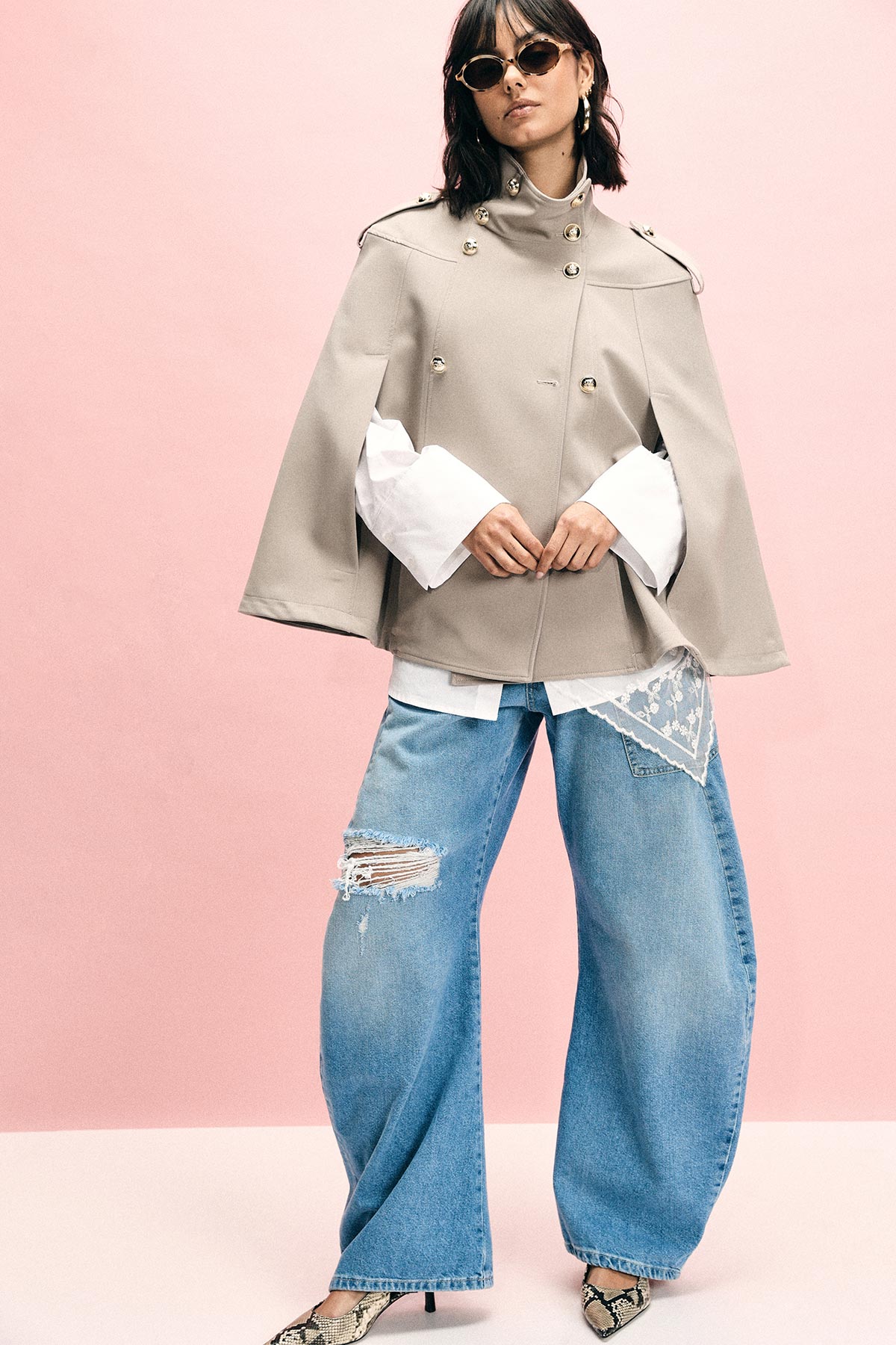 Woman in beige military-style cape over white shirt, wide-leg ripped jeans with lace patch, sunglasses and snake-print heels against pink backdrop