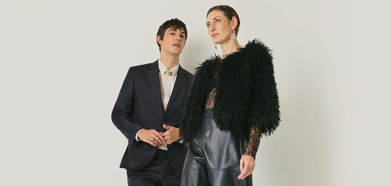 Due modelli posano su uno sfondo neutro; uno indossa un vestito scuro, mentre l'altro indossa una giacca di pelliccia ecologica nera, un top in pizzo, una gonna di pelle e stivali con il tacco.