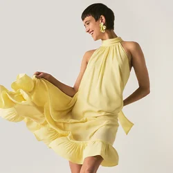 Persona in abito giallo e scarpe nere in piedi vicino a un ventilatore metallico da pavimento che soffia sull'abito, in una stanza semplice e neutra.