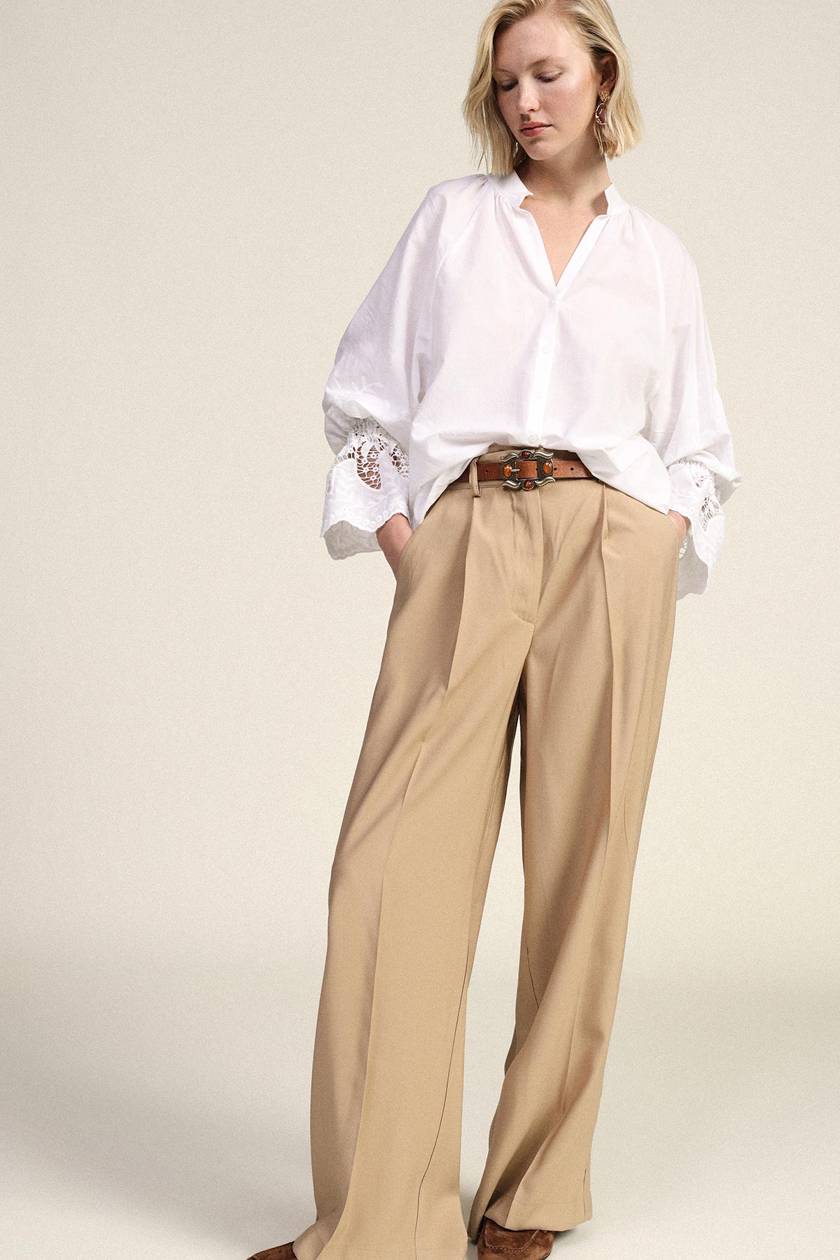Woman wearing an oversized white blouse with embroidered cuffs, high-waisted beige wide-leg trousers and a brown belt