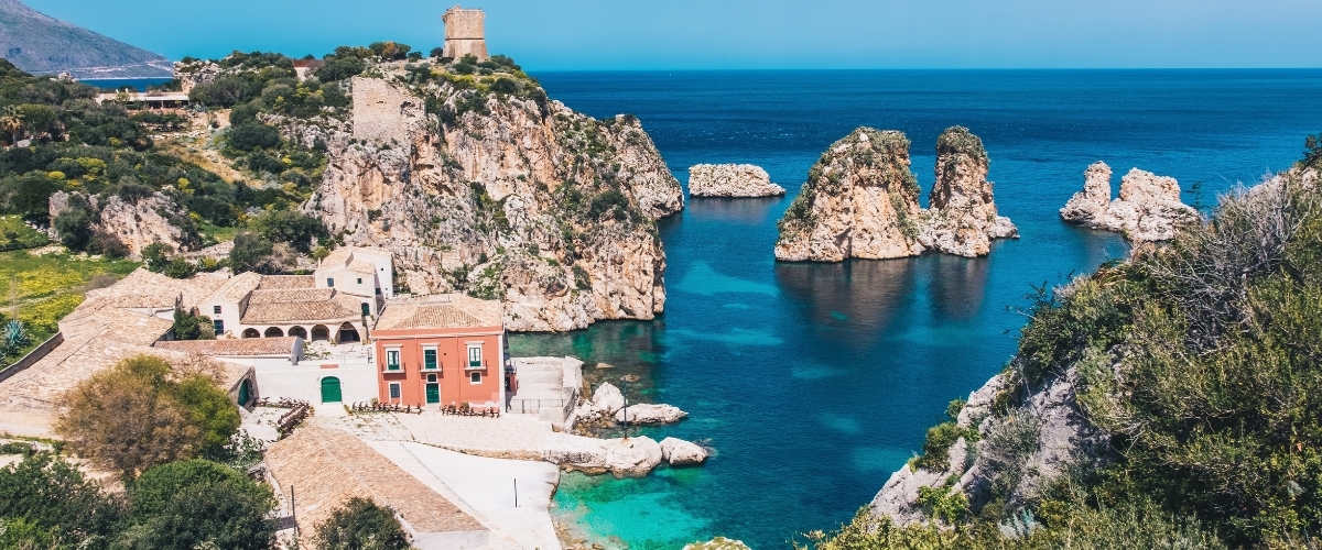 Aerial view of a Mediterranean cove with turquoise water, rocky sea stacks, cliffs, a historic stone tower and colourful coastal houses