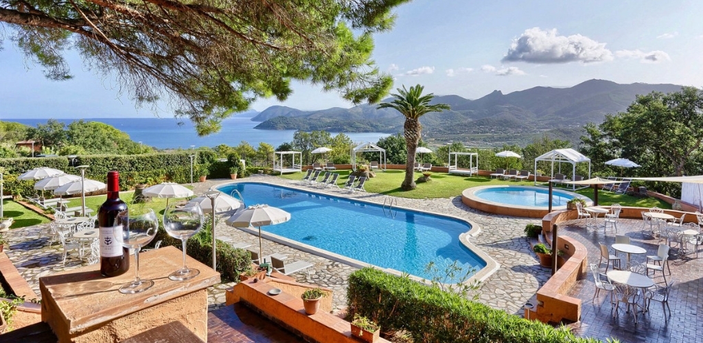 Resort pool terrace with bottle of red wine and glasses, sun loungers, cabanas and palm tree overlooking Mediterranean sea and mountains