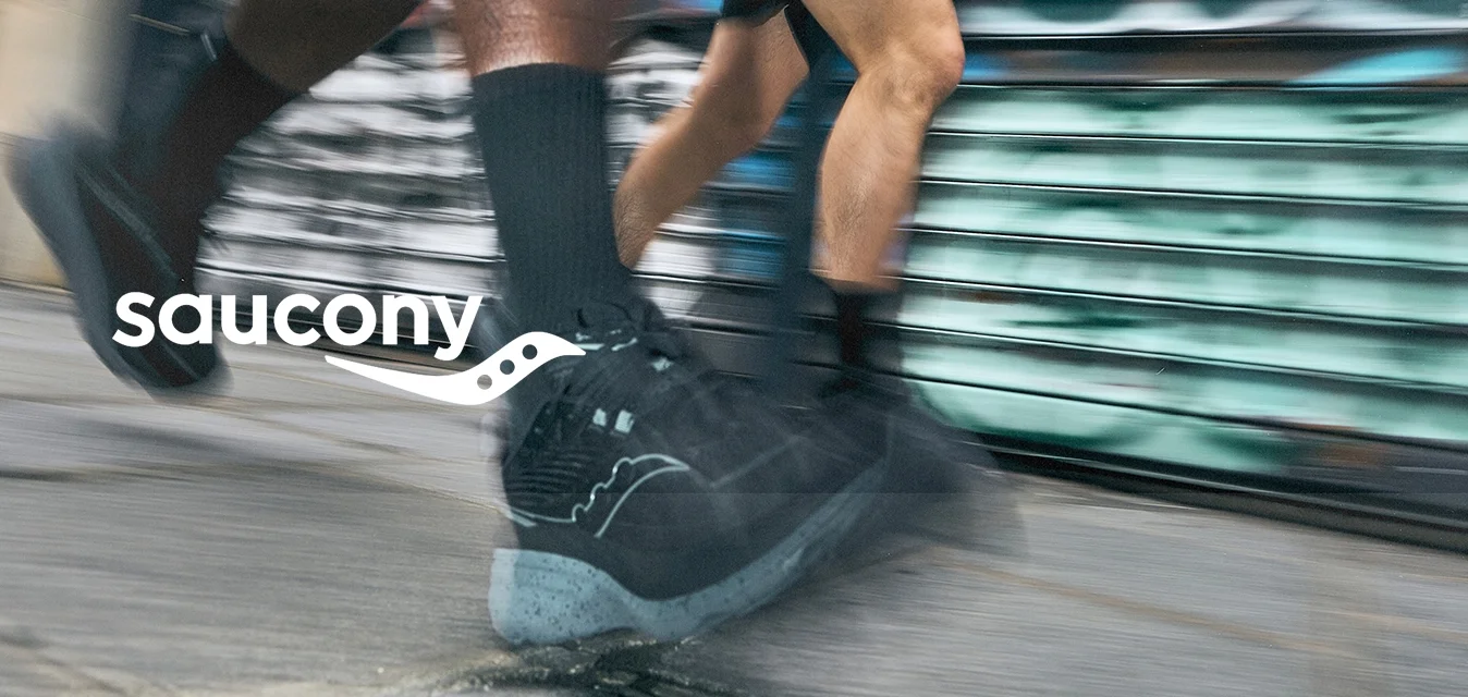 Scarpe sportive nere con una struttura testurizzata e suole grigie chiare in movimento su un pavimento, con uno sfondo urbano di graffiti sfocati.