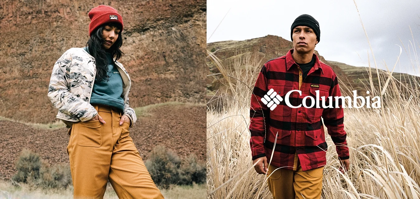 Giovane donna con berretto rosso, giacca floreale, top turchese e pantaloni senape in piedi su un terreno roccioso; uomo con berretto nero e giacca a quadri rossa nell'erba secca.