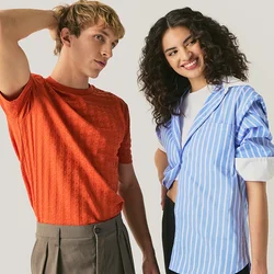 Giovane uomo con camicia arancione e pantaloni marroni in piedi con la mano sulla testa, donna con camicia a righe blu che sorride con la mano sui fianchi, sfondo neutro.