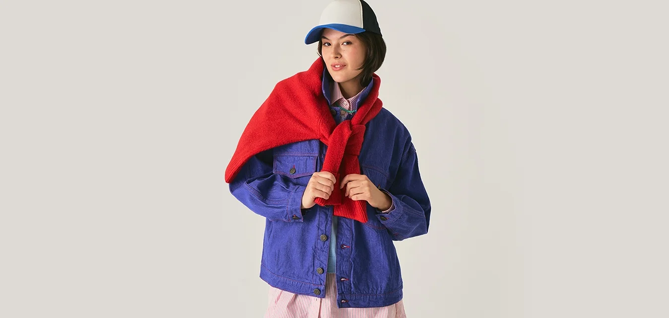 Giovane donna con una giacca blu e una sciarpa rossa drappata sulle spalle, indossa un cappello da baseball bianco e blu e si trova in un ambiente neutro.