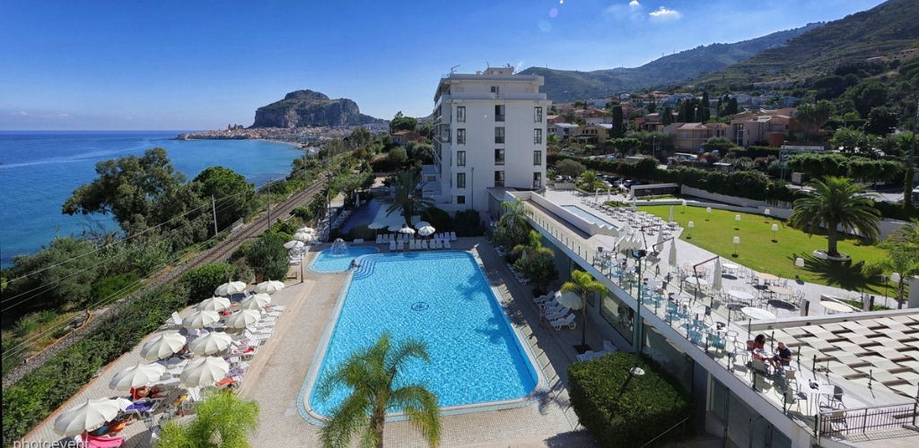 Seaside hotel with large outdoor swimming pool, sunbeds and umbrellas, palm trees, coastal railway and rocky headland under a clear blue sky