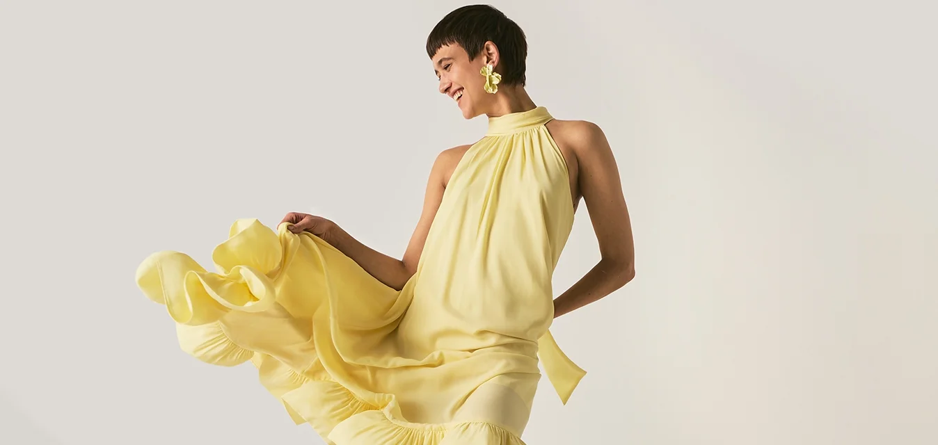 Persona in abito giallo e scarpe nere in piedi vicino a un ventilatore metallico da pavimento che soffia sull'abito, in una stanza semplice e neutra.