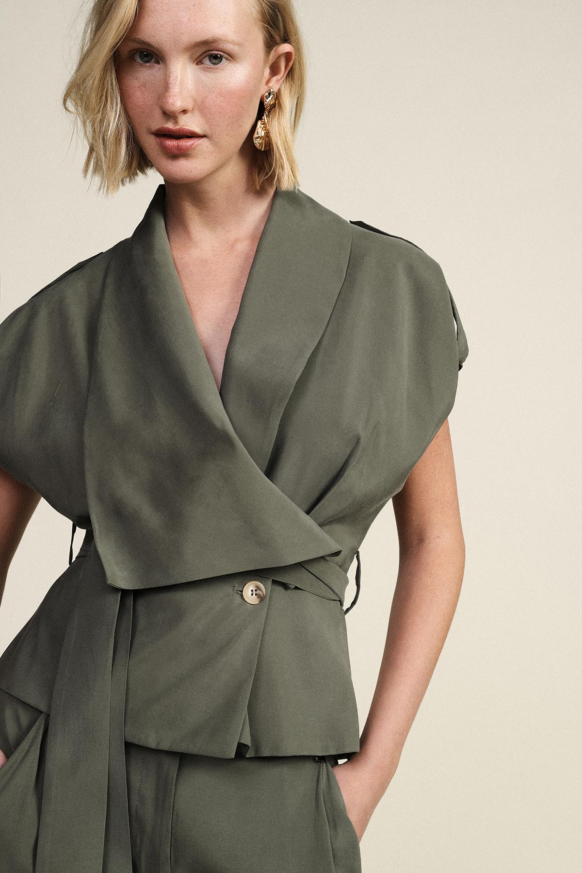 Model wearing olive-green wrap jacket and matching trousers, gold drop earrings on neutral background