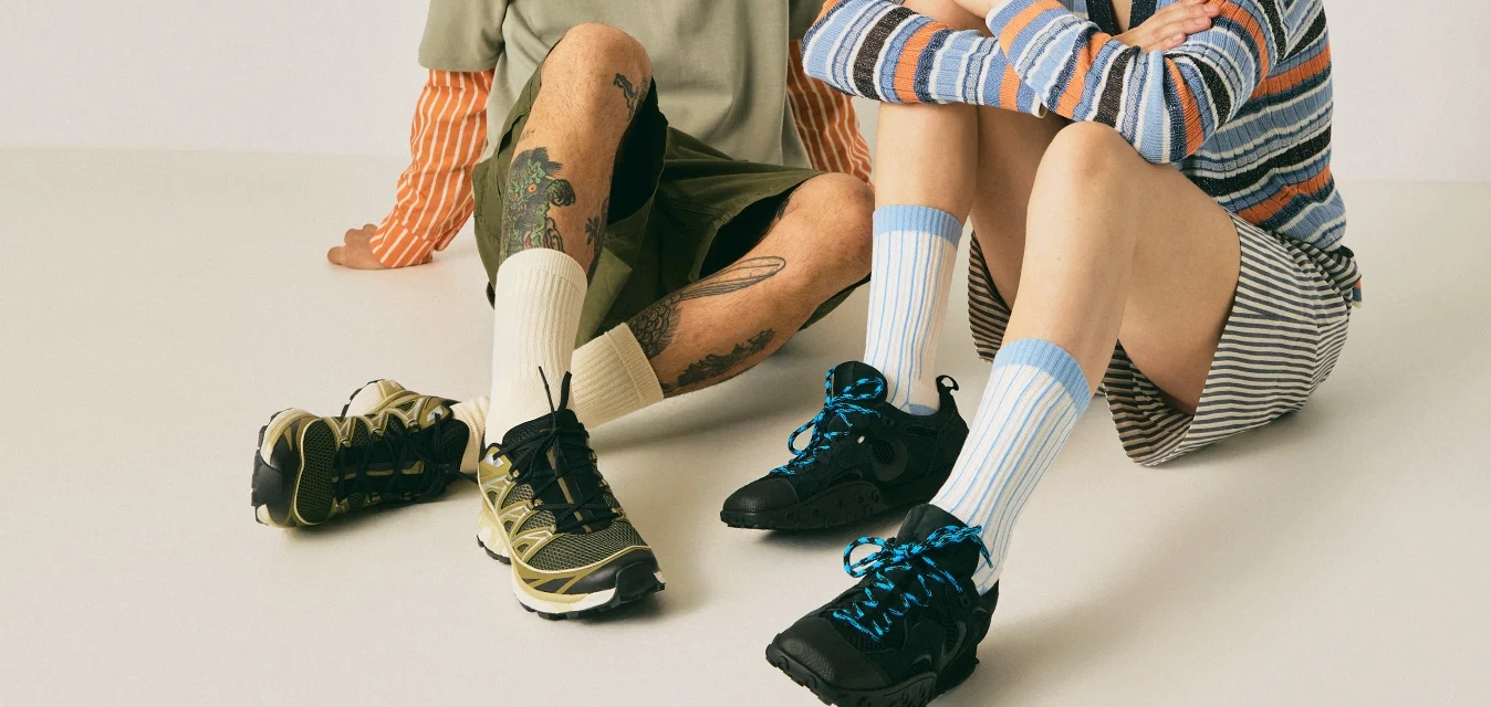 Due persone sedute sul pavimento contro uno sfondo semplice, vestite con abiti casual a righe e uniti, con sneakers e calze.