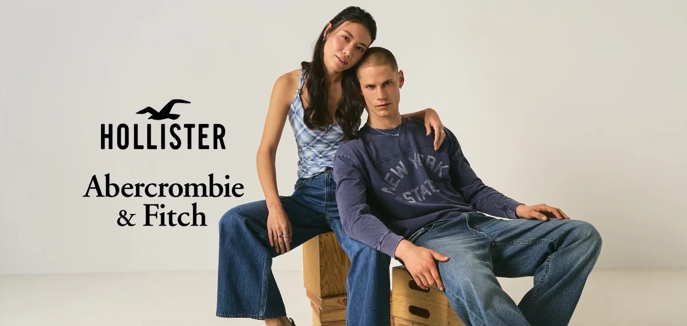 Giovane donna con canottiera e jeans larghi seduta su una cassa di legno con il braccio intorno a un giovane uomo con camicia a maniche lunghe e jeans seduto accanto a lei.