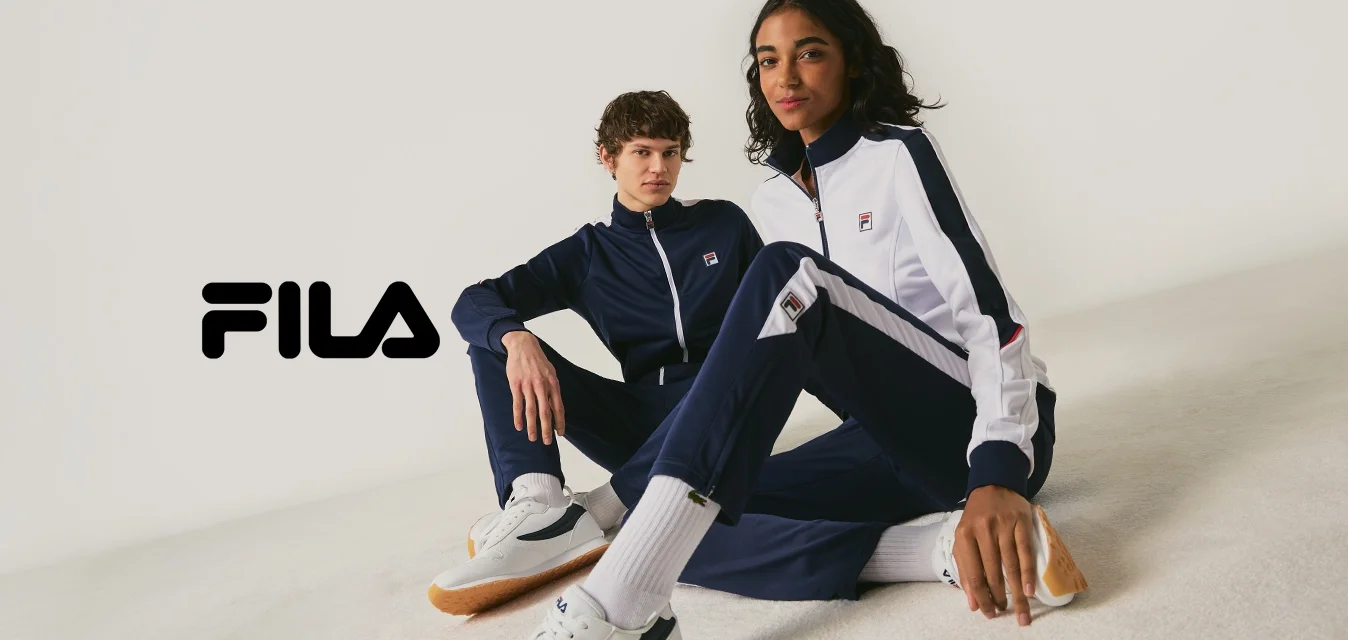 Due persone vestite con tute FILA blu navy e bianche e scarpe da ginnastica bianche, sedute sul pavimento contro uno sfondo chiaro e semplice.