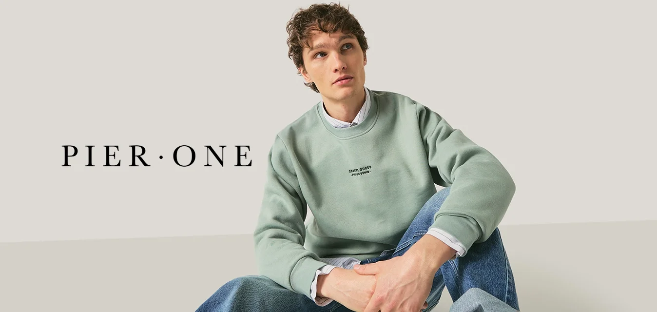 Giovane con capelli ricci seduto a gambe incrociate, indossando una felpa verde chiaro e jeans blu risvoltati, su uno sfondo semplice.