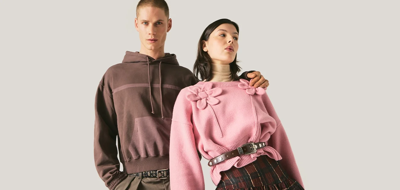 Due modelli stanno affiancati; uno indossa una felpa con cappuccio e pantaloni marroni, l'altro un maglione rosa con dettagli floreali e una gonna a quadri.