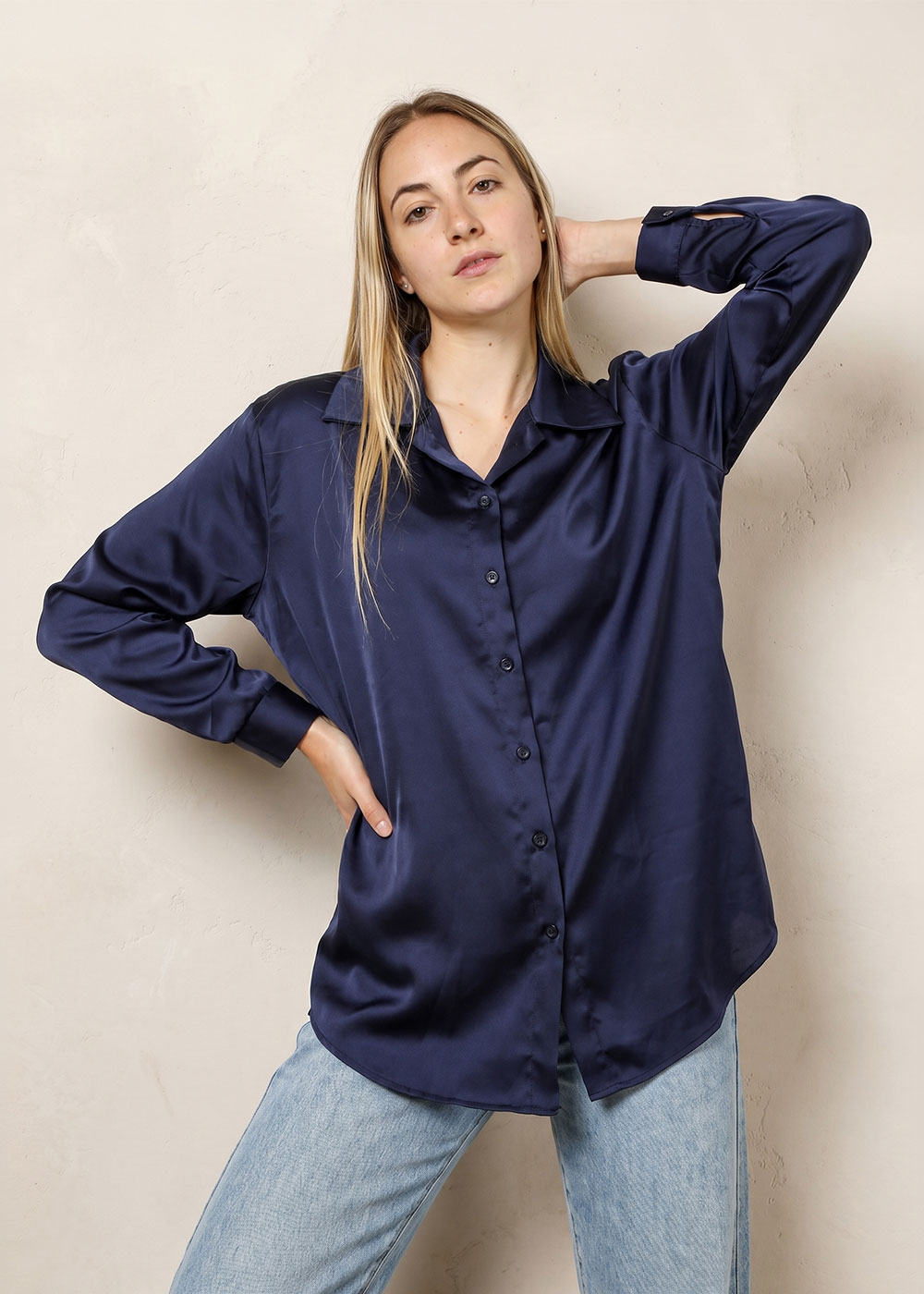 Woman in a navy satin button-up blouse and light blue jeans posing against a neutral textured wall