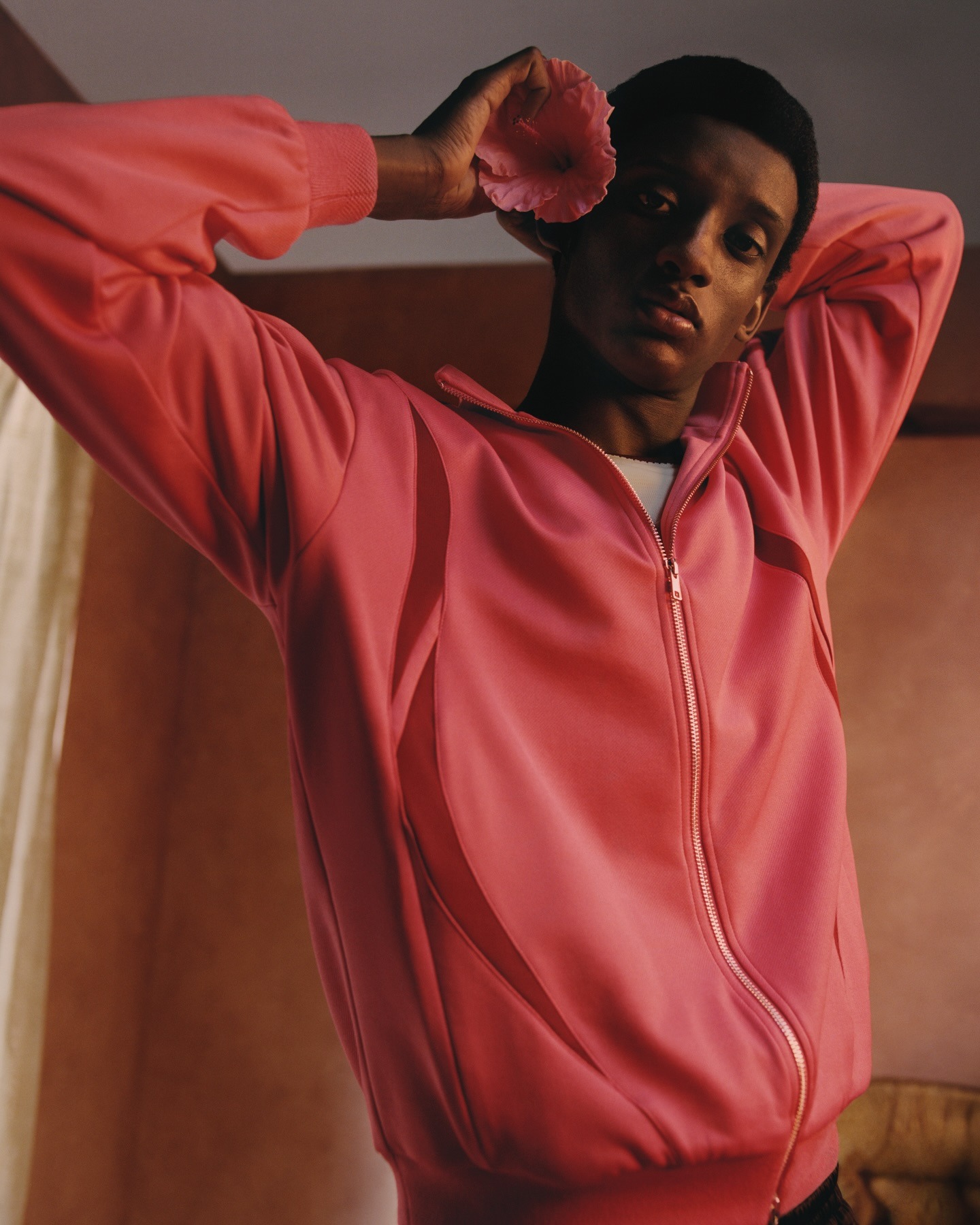 Portrait of model in a pink zip-up jacket holding a pink flower to their ear against a warm backdrop