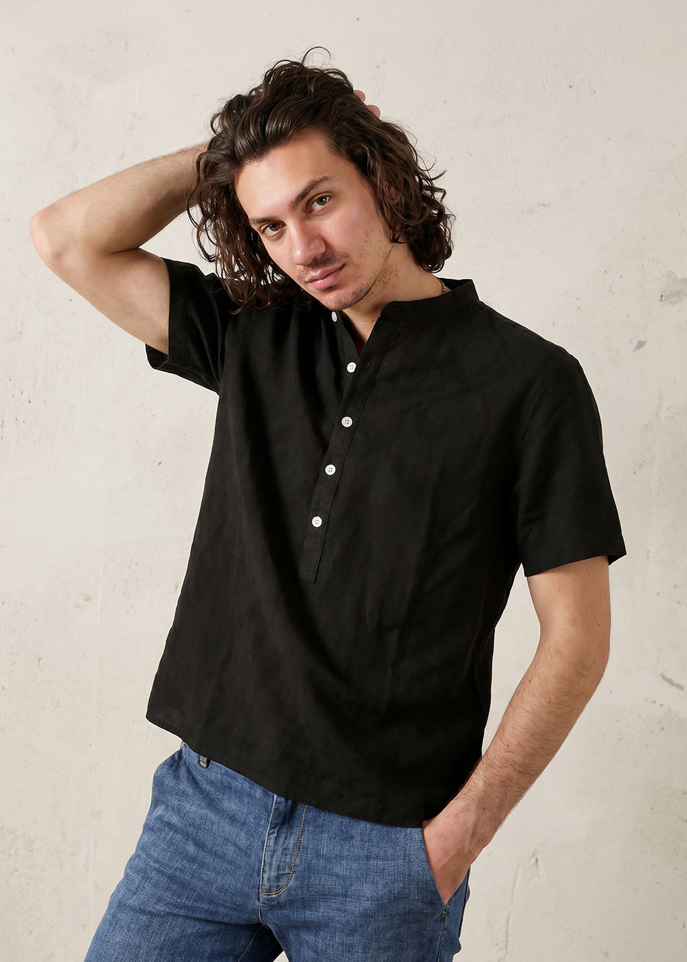 Young man wearing a black short-sleeve mandarin-collar shirt and blue jeans, one hand in pocket and the other touching his hair against a neutral background