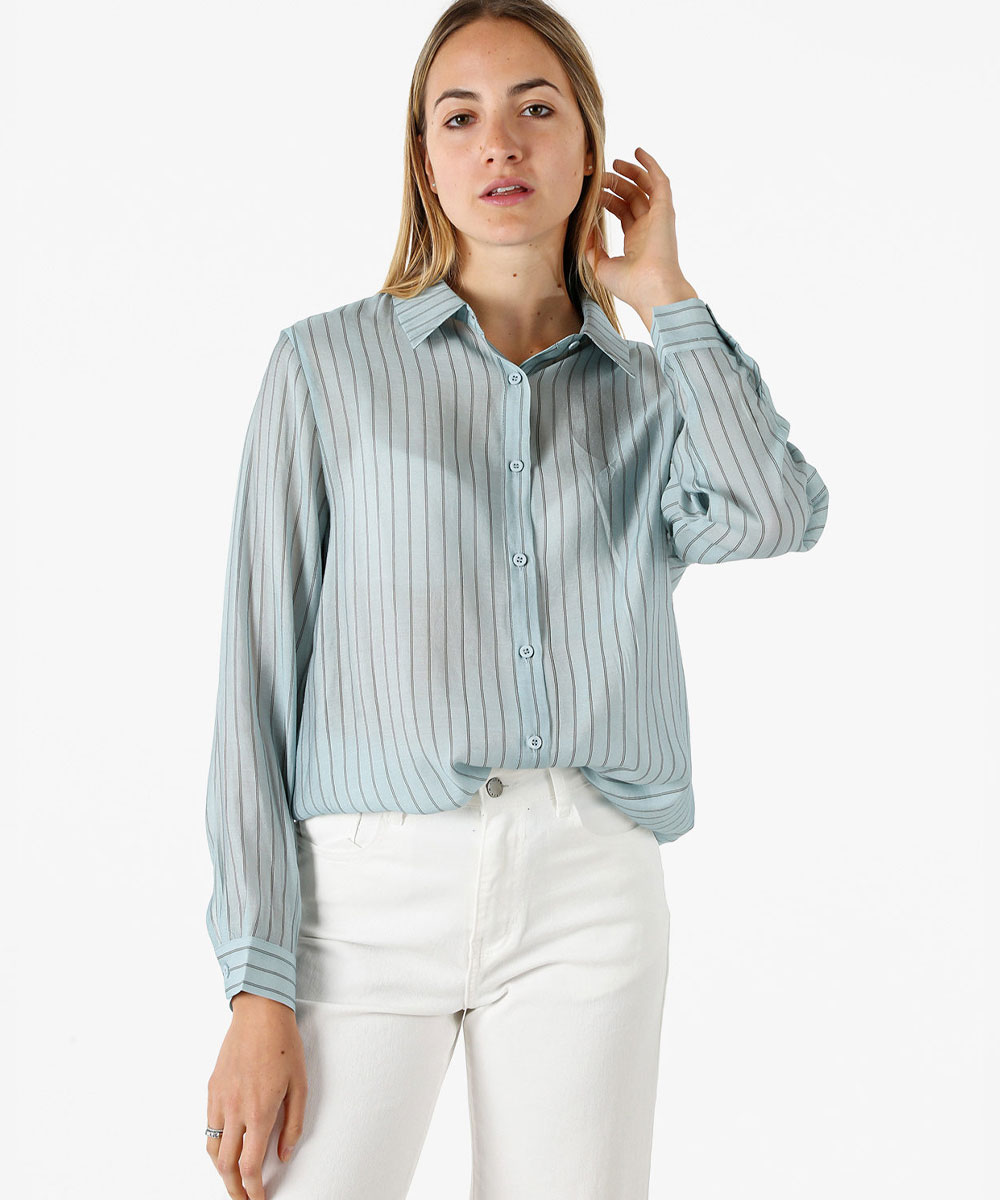 Model wearing a light-blue pinstripe long-sleeve button-up blouse with white high-waisted jeans, hand near hair, on white background