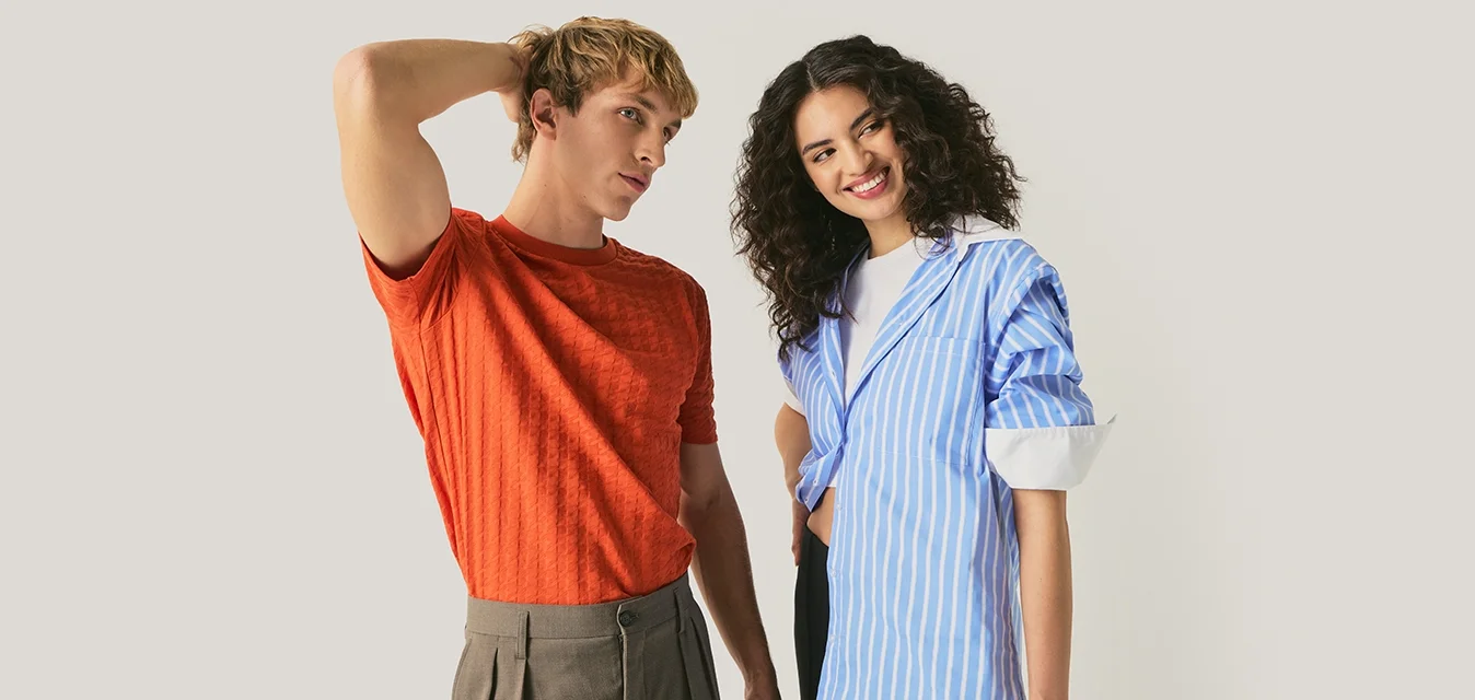 Giovane uomo con camicia arancione e pantaloni marroni in piedi con la mano sulla testa, donna con camicia a righe blu che sorride con la mano sui fianchi, sfondo neutro.