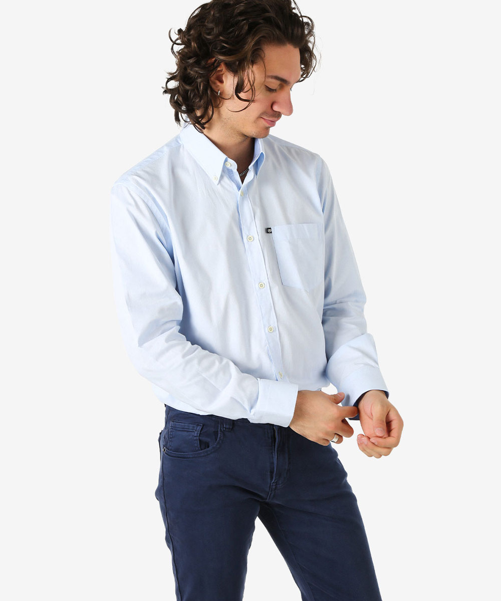 Male model adjusting cuff of light blue button-down shirt paired with navy chinos on white background