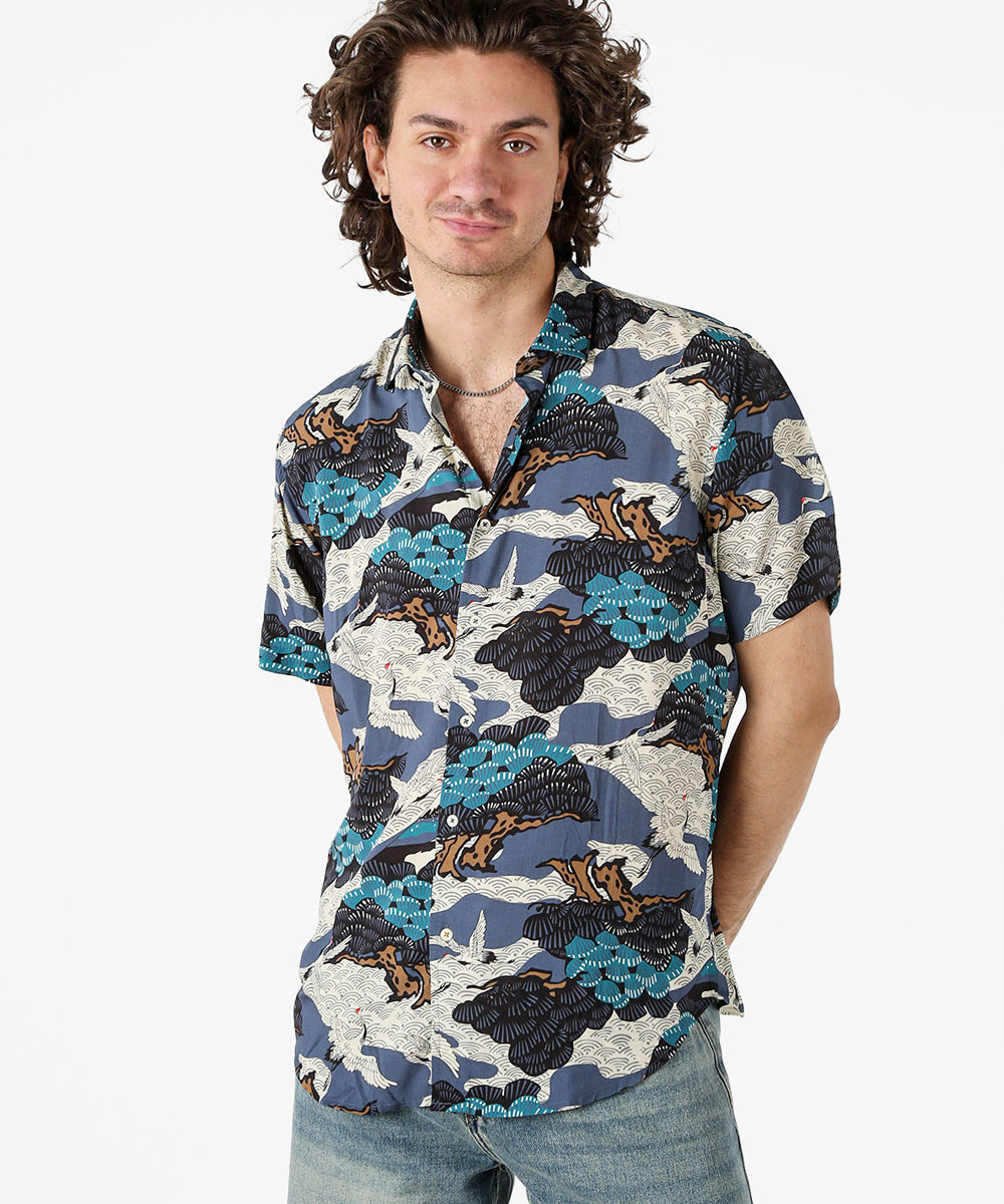 Man wearing a short-sleeved button-up shirt with blue, teal and beige wave and cloud print, paired with jeans