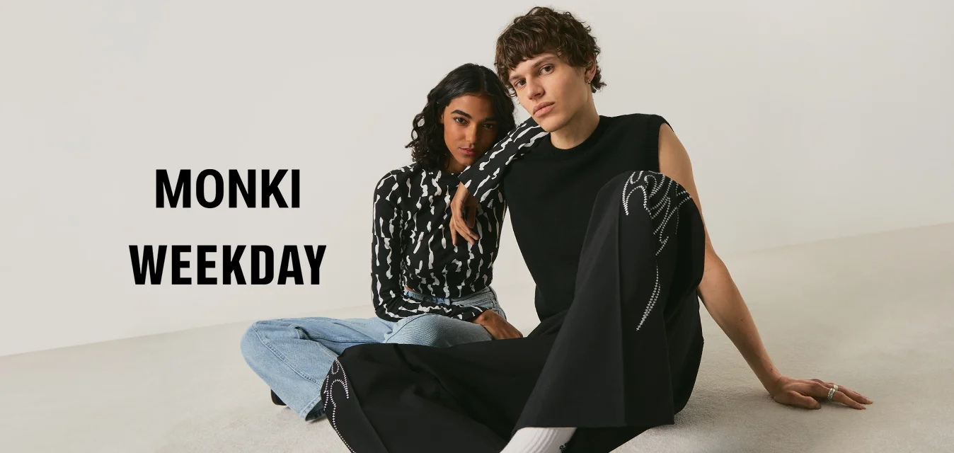 Due giovani adulti seduti a terra contro una parete bianca; uno con top nero senza maniche e pantaloni, l'altro con maglione con motivo bianco e nero e jeans.