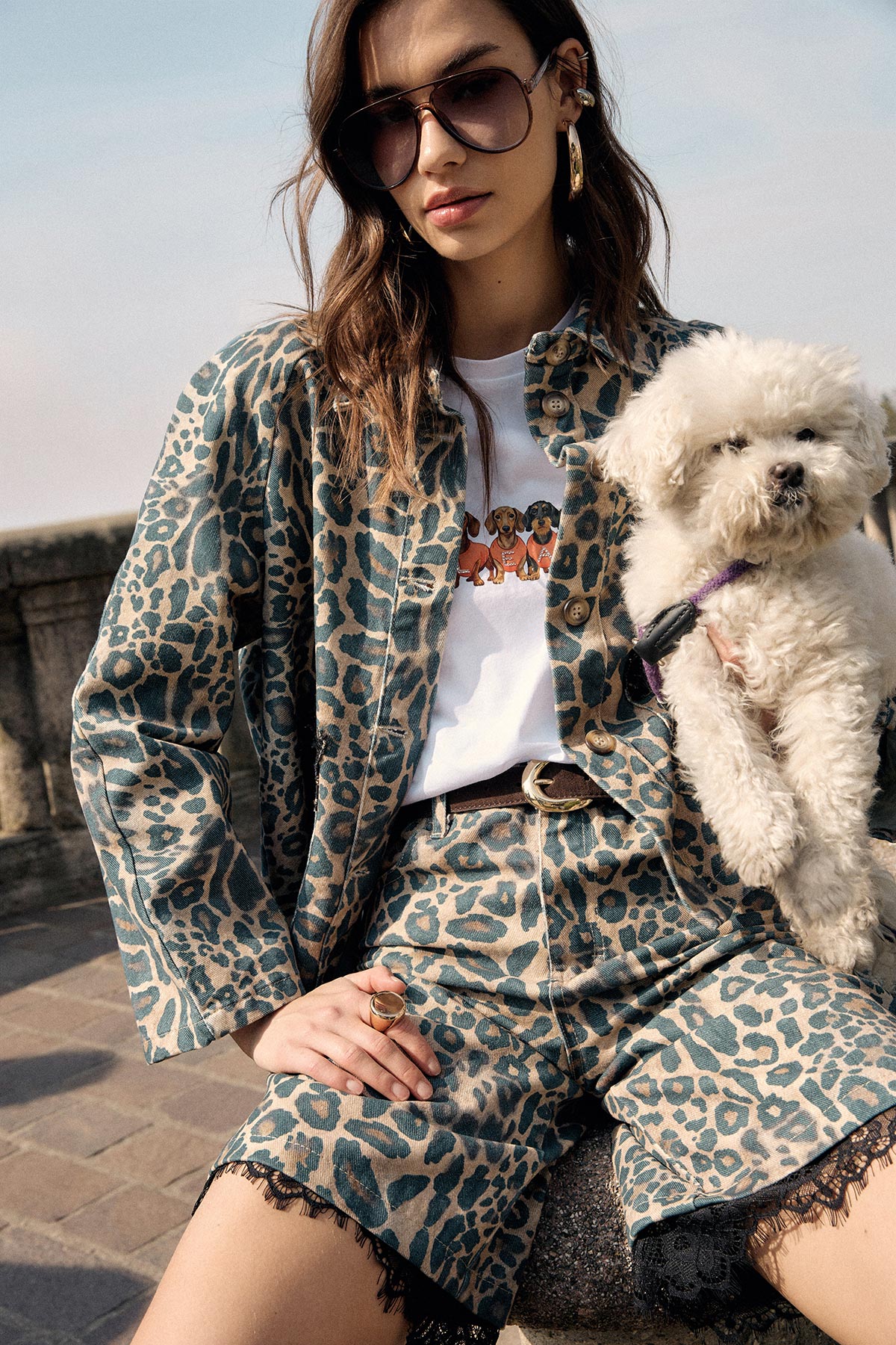 Woman in leopard-print jacket and shorts wearing sunglasses, seated on a stone bench holding a small white fluffy dog