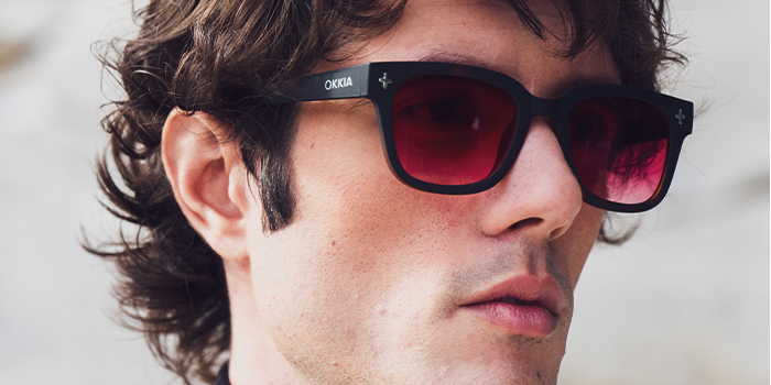 Close-up of a young man wearing black-frame sunglasses with red lenses and visible OKKIA logo
