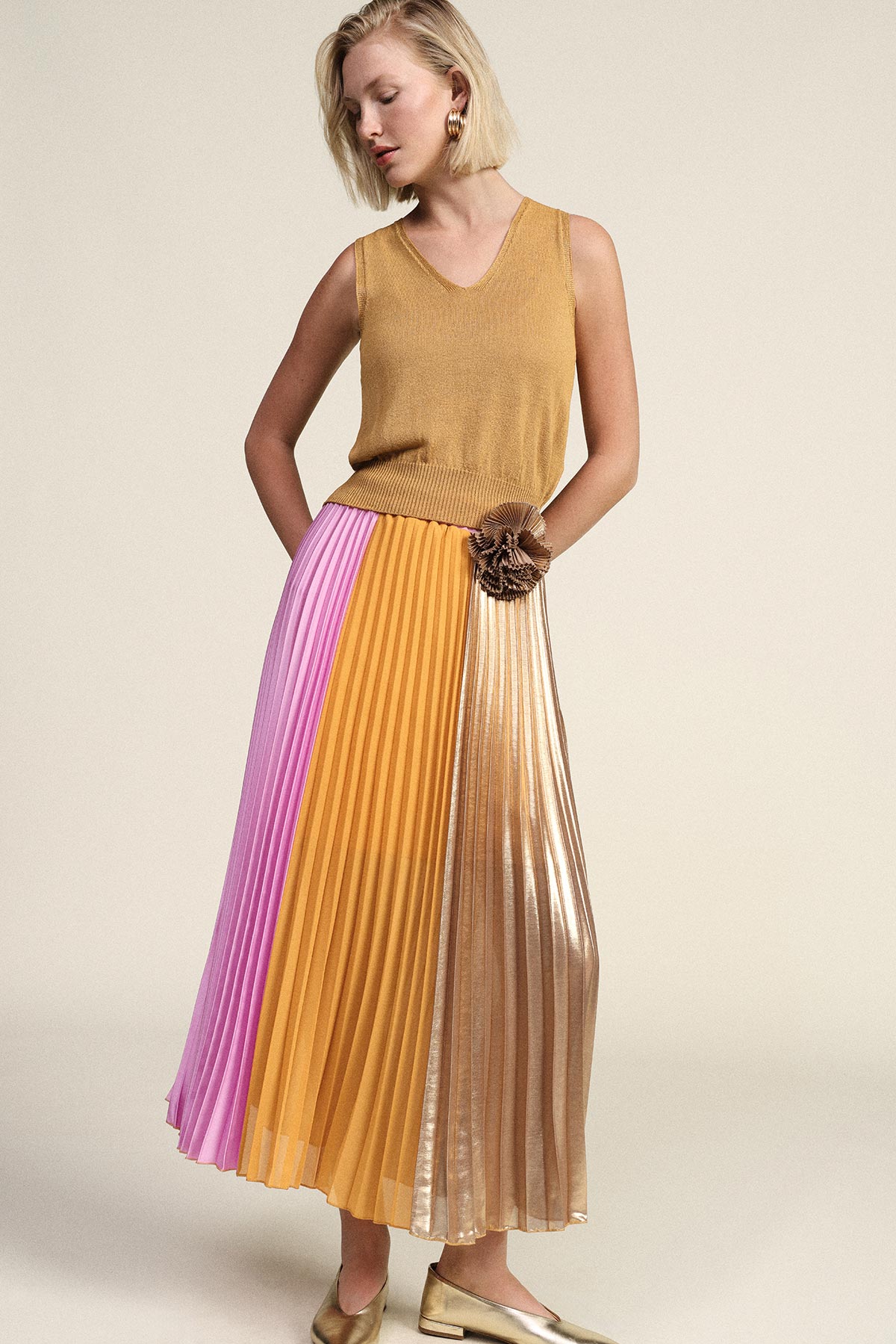Woman in sleeveless mustard top and colour-block pleated midi skirt (pink, ochre, metallic gold) with gold flats, hands in pockets