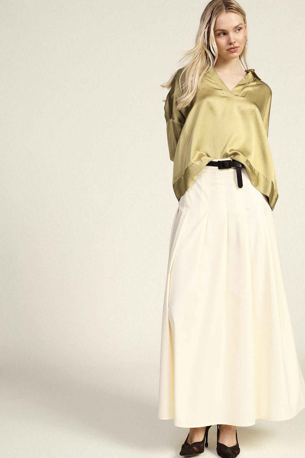 Female model wearing an olive satin blouse tucked into a high-waisted cream maxi skirt with a black belt and heels, standing against a pale background