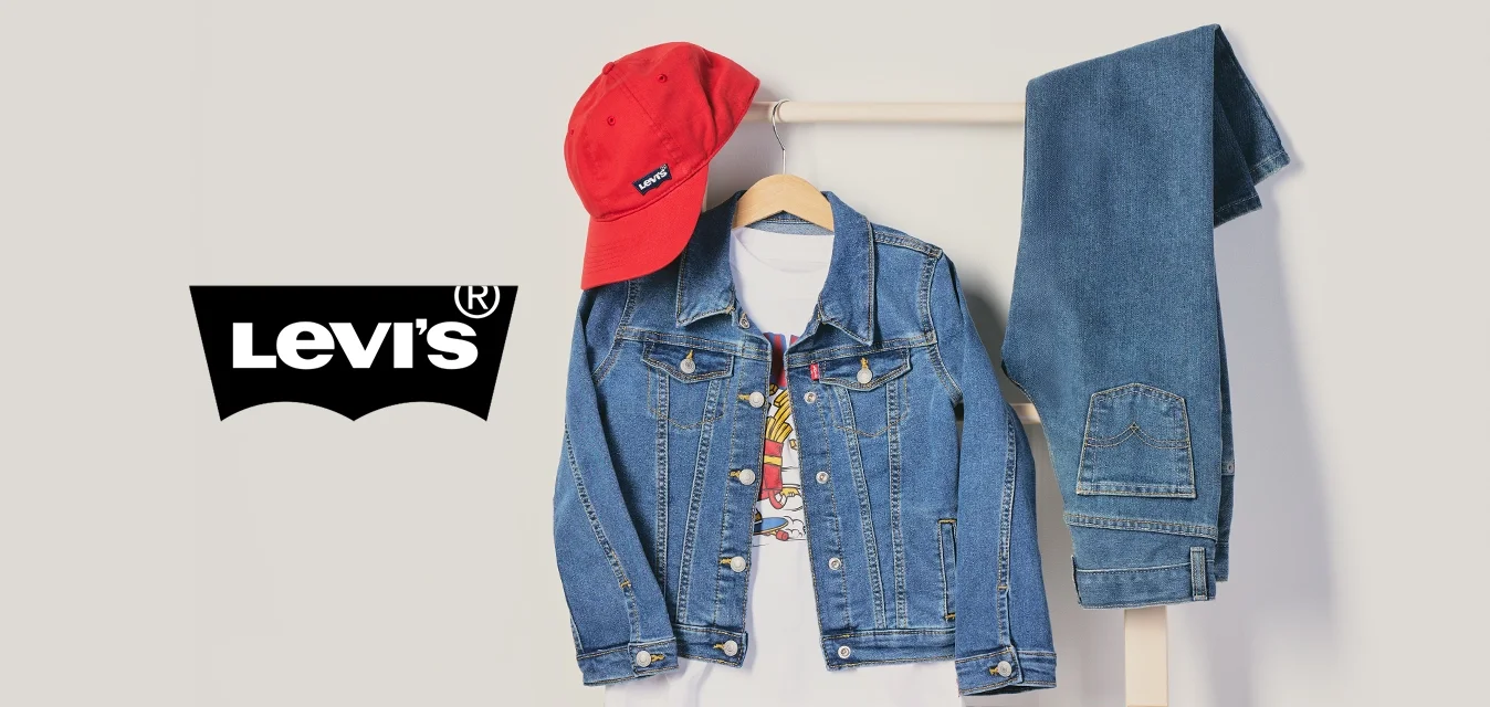 Cappellino rosso, giacca di jeans sopra una t-shirt bianca e jeans blu piegati appesi a un appendiabiti in legno su uno sfondo semplice.