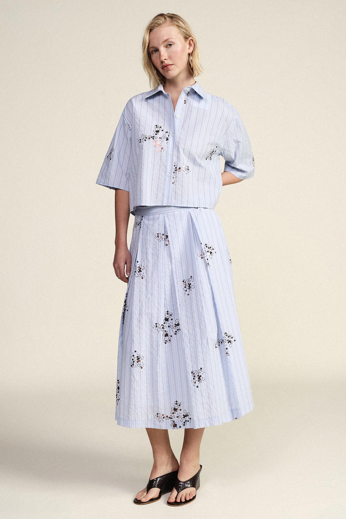 Woman wearing light-blue striped two-piece set with button-up cropped shirt and pleated floral midi skirt, black sandals