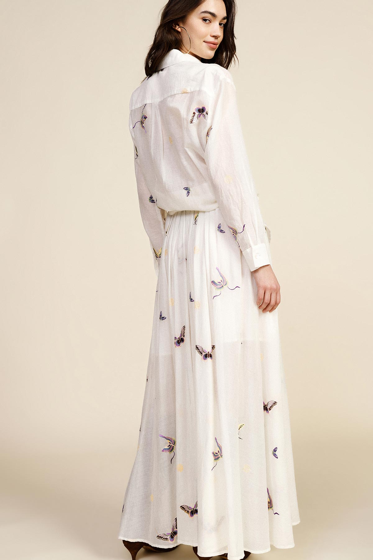Woman wearing white embroidered long-sleeve maxi dress, looking over shoulder, pleated skirt on neutral studio background