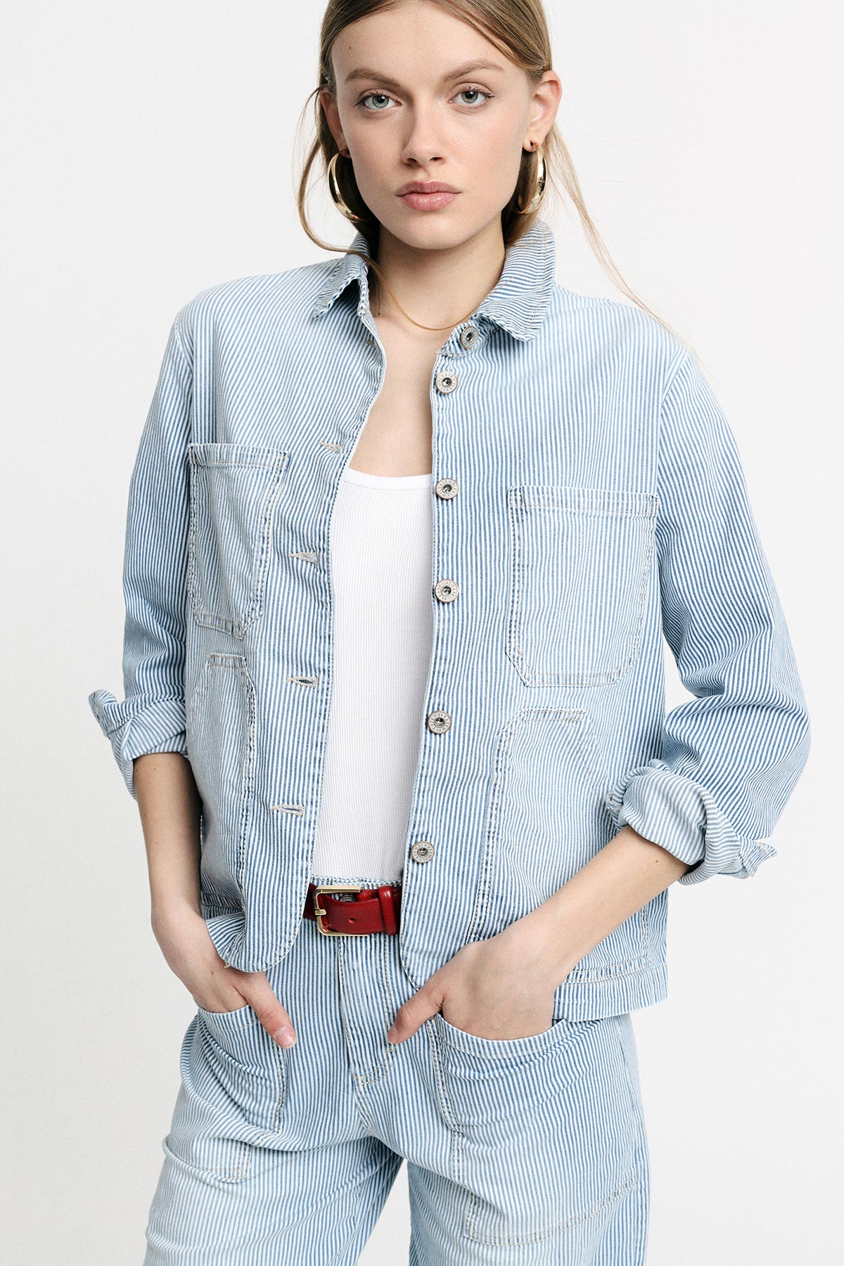 Young woman wearing light-blue pinstripe jacket and matching trousers, white vest top, red belt and gold hoop earrings, hands in pockets