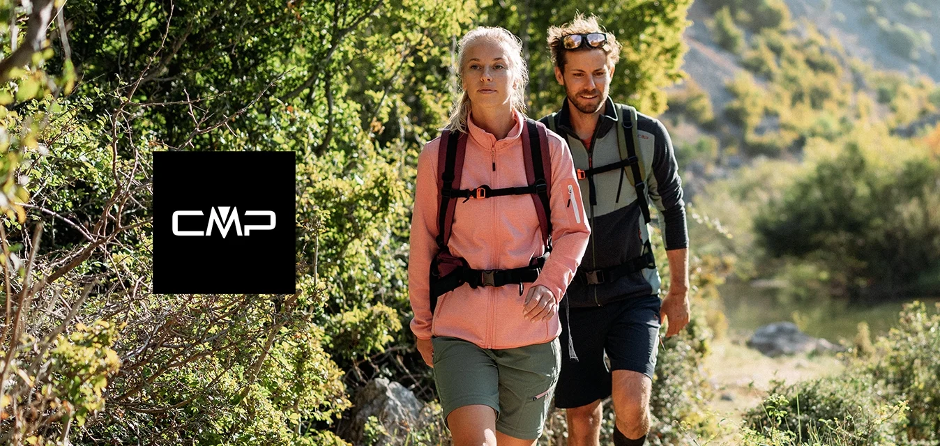 Due escursionisti con abbigliamento da esterni camminano su un sentiero sterrato accanto a un fiume, circondati da alberi verdi e colline illuminate dal sole.