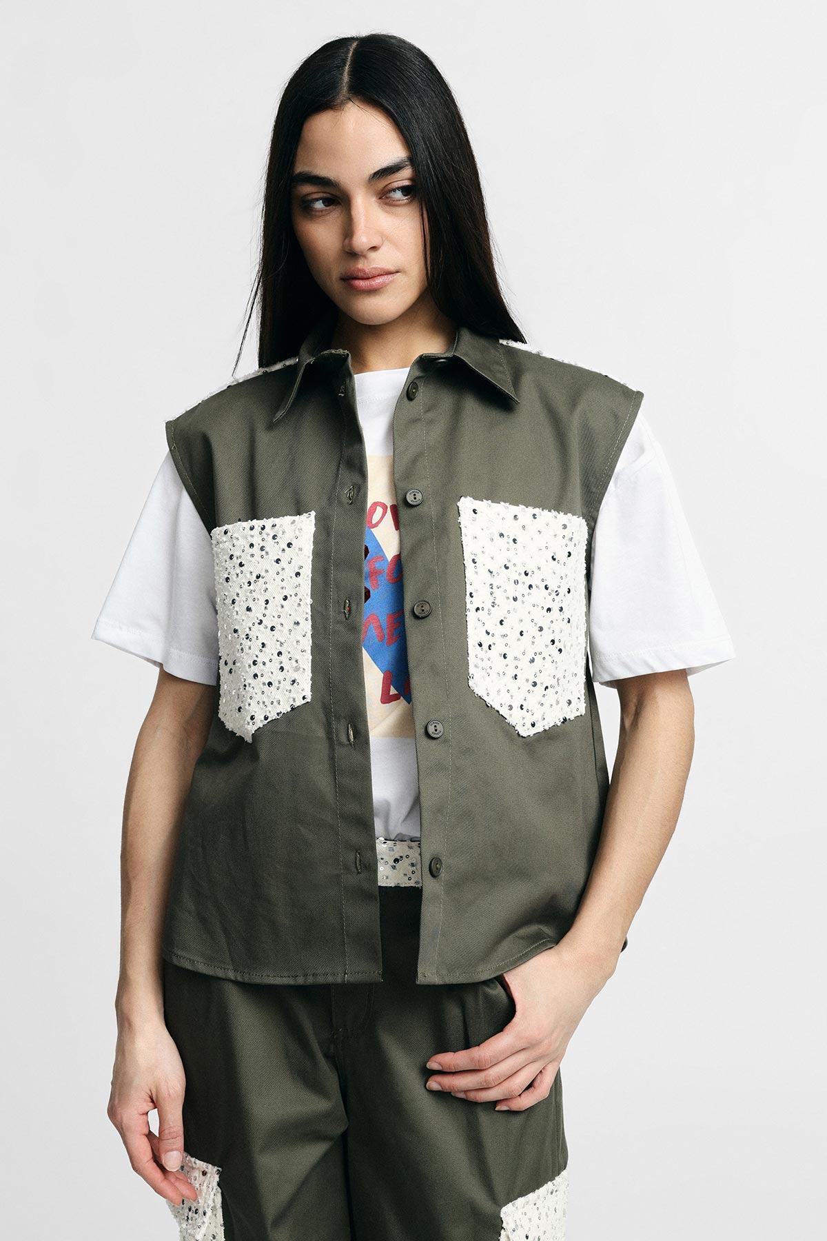 Model wearing olive-green sleeveless jacket with white patterned patch pockets, matching shorts and white graphic t-shirt