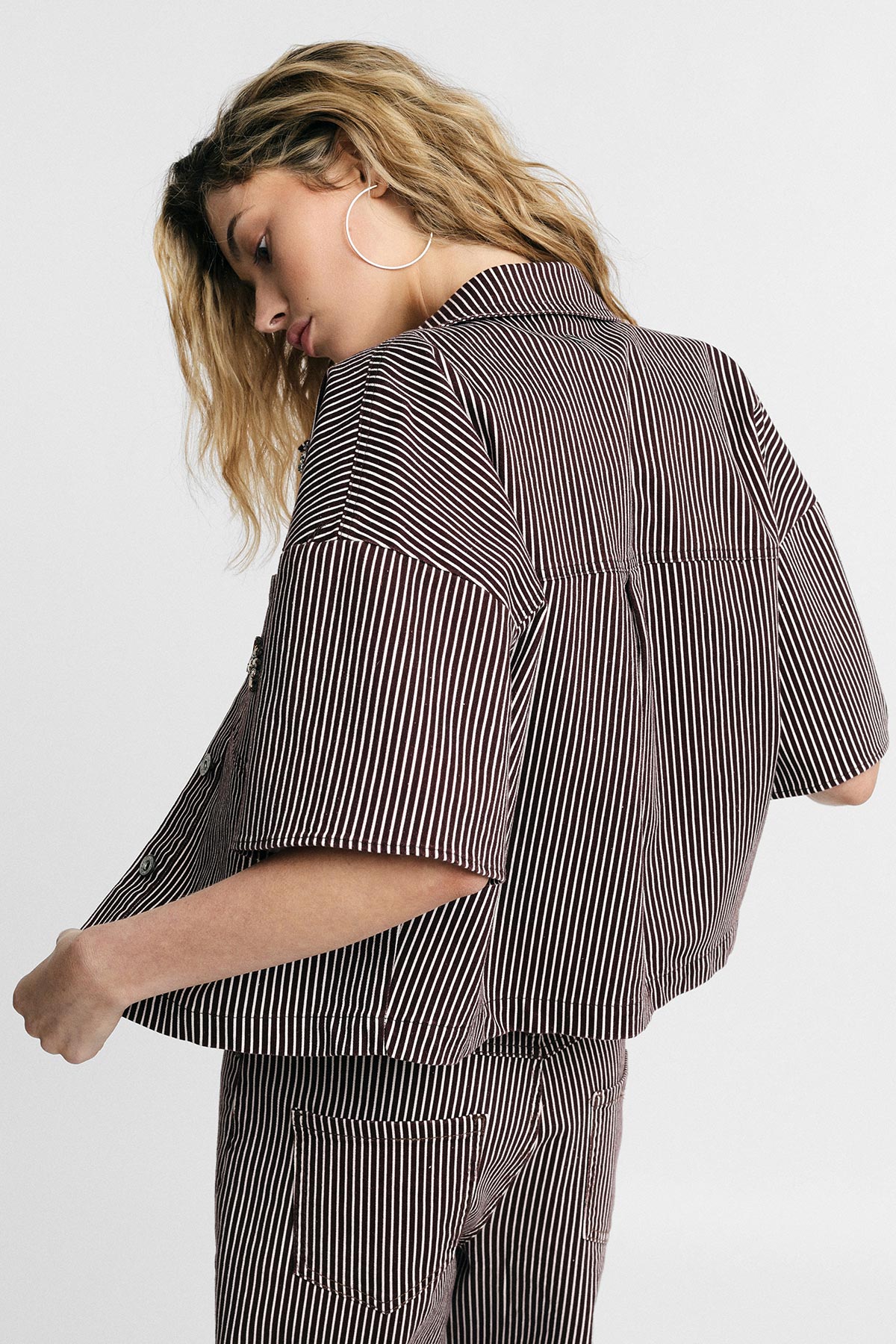 Woman in brown-and-white striped short-sleeve jacket and matching trousers, large hoop earring, side-back pose against plain background
