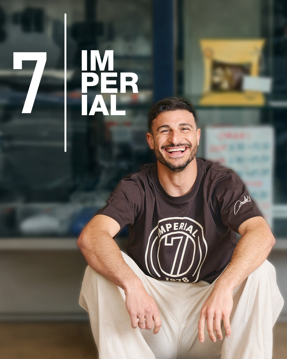 Smiling man wearing an Imperial 7 logo t-shirt, sitting casually outside a shop-front window, relaxed urban portrait