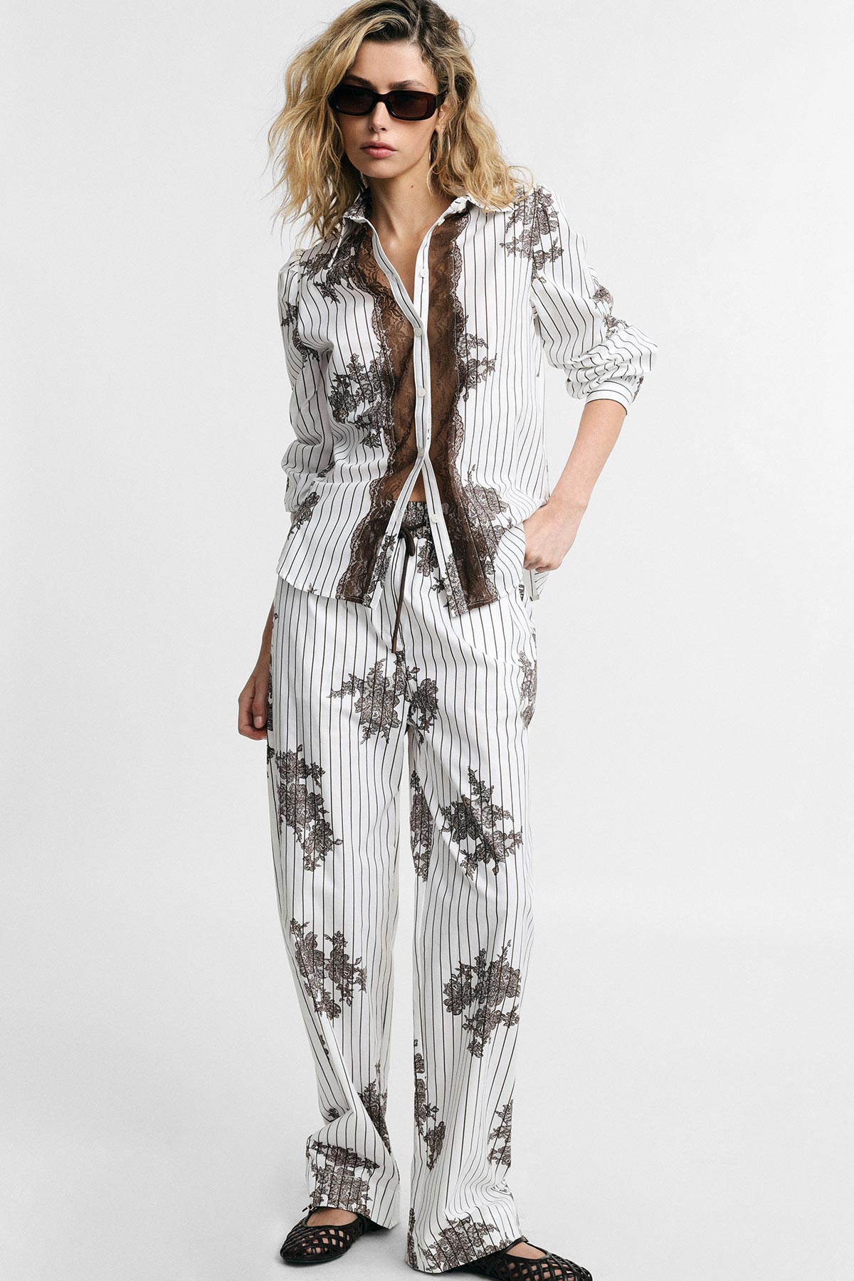 Female model wearing white striped pyjama-style suit with brown floral print, buttoned shirt, wide-leg trousers and black sunglasses