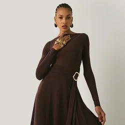 Abito lungo marrone con maniche a lunghezza completa e design avvolgente, caratterizzato da una fascia laterale e un accento a forma di anello dorato in vita. Tessuto morbido con una silhouette fluida.