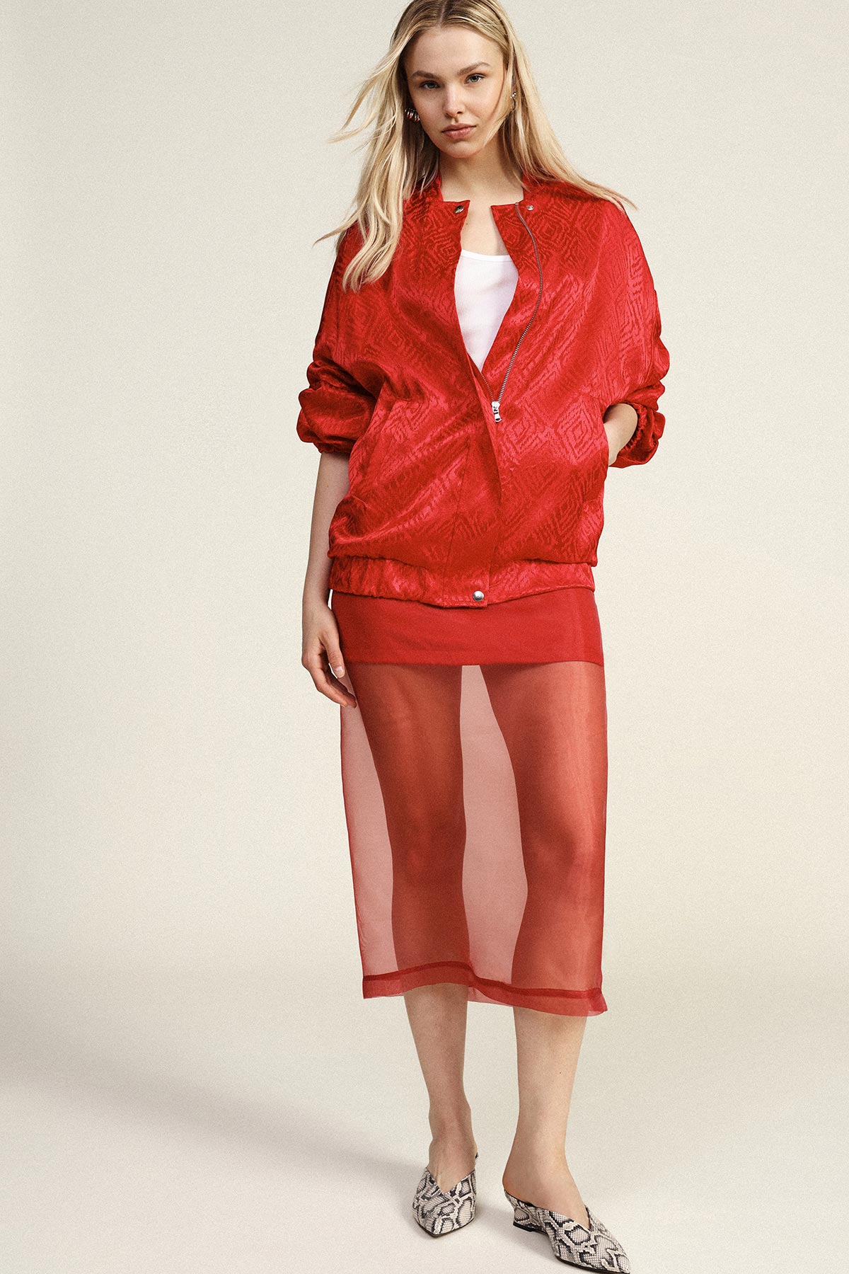 Woman in red textured bomber jacket, white top and sheer red midi skirt with snakeskin mules, studio fashion pose