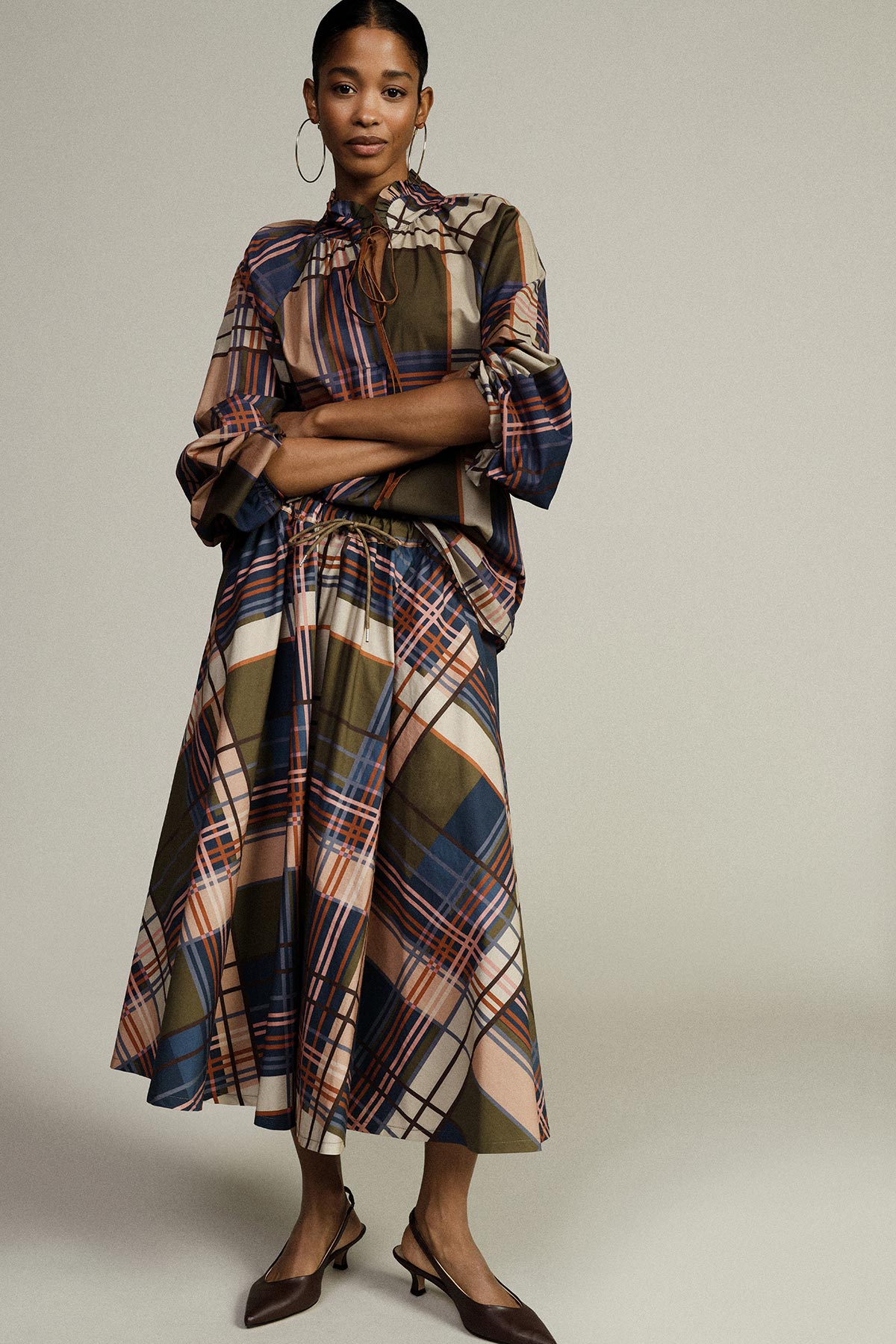Woman in matching oversized multicolour plaid blouse and A-line midi skirt, hoop earrings and brown slingback heels on neutral background