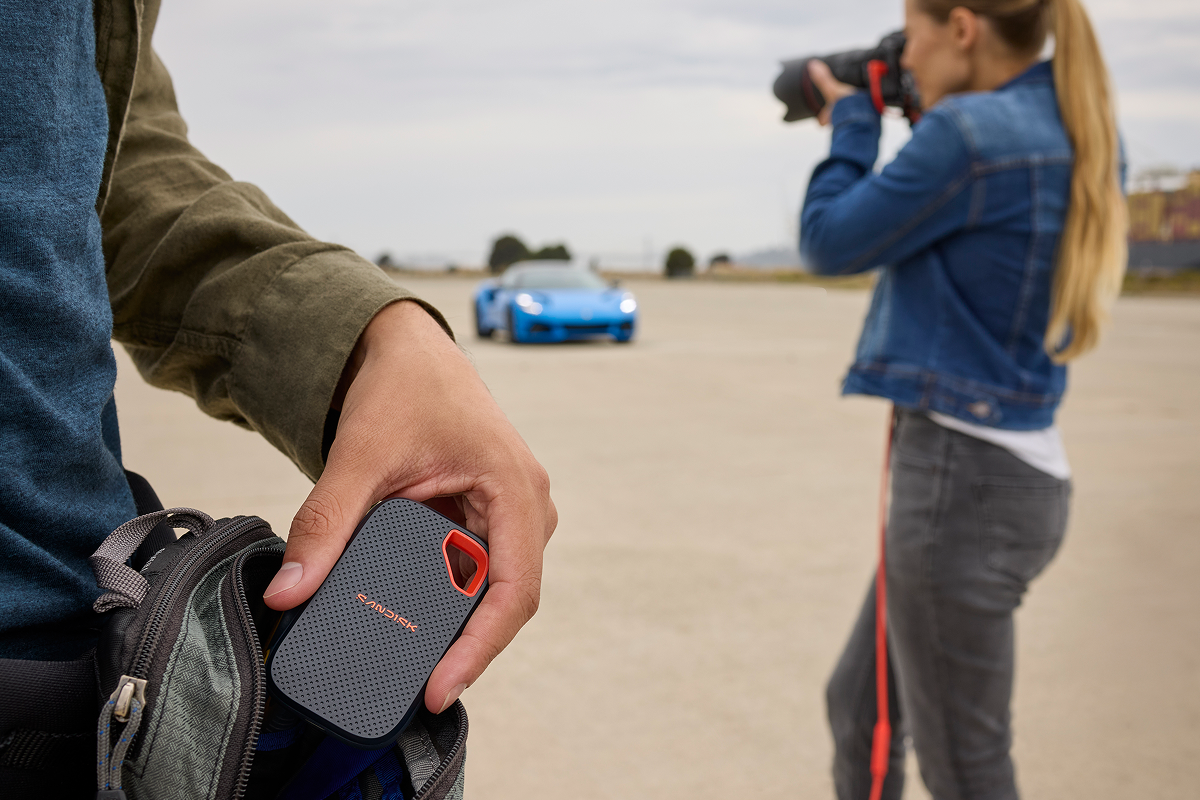 Una mano recupera un SSD portatile SANDISK Extreme&reg; da una borsa, con un fotografo che riprende un'auto sportiva blu sullo sfondo.